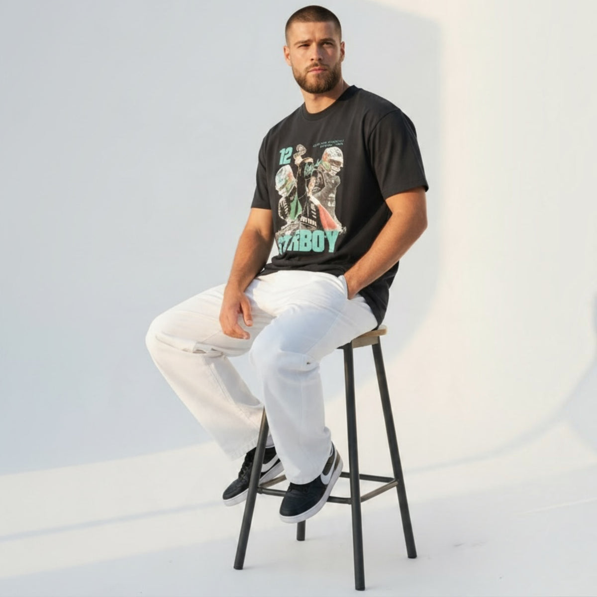 Modello indossa t-shirt nera Chulo Store in cotone con grafica motorsport, abbinata a pantaloni bianchi su sgabello